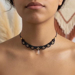 May include: A black macrame choker necklace with gold-colored beads and a teardrop-shaped pendant. The necklace is worn around the neck and is a close-up shot. The background is blurred.