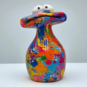 May include: A brightly coloured ceramic vase with a distinctive, abstract design. The vase has a rounded body and a flared top with two white, googly eyes. The surface is covered in vibrant paint splatters of blue, orange, yellow, and pink.