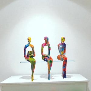 Op de afbeelding: Drie abstracte sculpturen van zittende figuren, elk met een uniek kleurrijk verfspatontwerp. De figuren zijn geplaatst op een doorzichtige glazen plank boven een witte plank met planten.