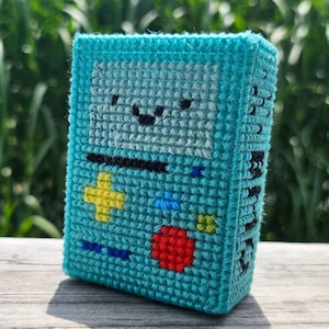 May include: A turquoise, pixelated, handmade BMO-inspired case. The case features a smiling face, a black bar, a yellow cross, a red circle, and other colorful details. The case is made of a textured material and sits on a wooden surface.