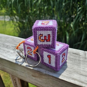 May include: Three purple and white handmade needlepoint blocks with the numbers 1, 2, and 3 in orange. The blocks are attached to a silver keychain with an orange cord. The blocks are sitting on a wooden surface.