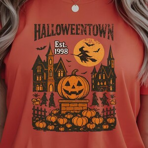 以下が含まれることがあります： ハロウィーンをテーマにしたグラフィックが特徴のオレンジ色のTシャツ。「Halloweentown Est. 1998」の文字と、カボチャ、魔女、お化け屋敷のイラストが描かれています。半袖のクルーネックTシャツです。