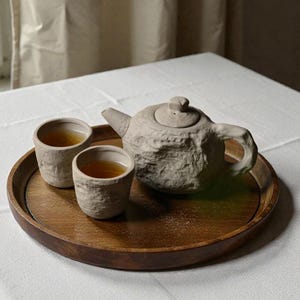 May include: A tea set featuring a textured, light gray teapot and two matching cups filled with tea, presented on a round wooden tray. The tray sits on a white tablecloth, with a window and curtains in the background.