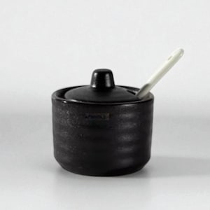 May include: A black ceramic sugar bowl with a lid and a small white spoon. The bowl has a textured surface and a small handle on the lid. The spoon is inserted into the bowl. The bowl is likely 8-10 cm in diameter.