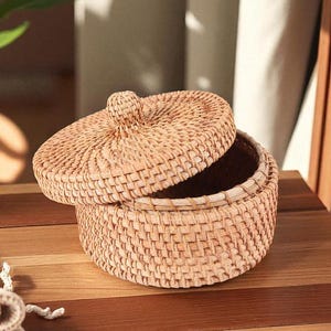 May include: A round, woven basket with a lid, made of light brown rattan. The lid is slightly ajar, revealing the interior of the basket. The basket sits on a wooden surface, with a plant and a folded cloth in the background.