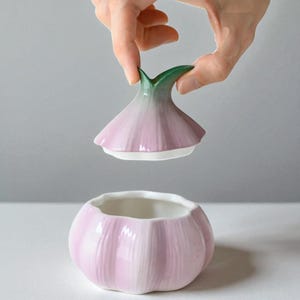 Puede incluir: Un recipiente de cerámica con forma de ajo con tapa extraíble. El recipiente y la tapa son de color rosa claro con detalles en blanco y verde. La tapa está siendo levantada por una mano. El recipiente está sobre una superficie blanca.