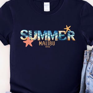 May include: Navy blue t-shirt with the word "SUMMER" in a colorful design, featuring ocean waves and starfish. Below the word "SUMMER" is the word "MALIBU" and the year "2026". The shirt is paired with blue jeans.