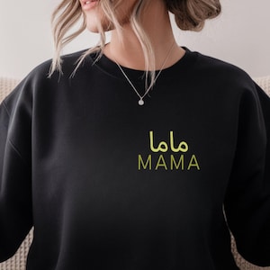 May include: Black sweatshirt with the word "MAMA" in both Arabic and English, in a light green color. The sweatshirt is a crew neck style and appears to be made of a soft material. The design is located on the upper left chest area.