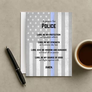 May include: A greeting card with the American flag design and the words "A prayer for POLICE." The card includes a prayer for protection, strength, wisdom, and guidance. A black pen and a cup of coffee are on the table.