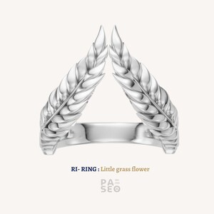 Puede incluir: Un anillo plateado con un diseño de dos espigas de trigo estilizadas que forman un triángulo. El anillo está etiquetado como "RI- RING : Little grass flower" en texto azul. El anillo está sobre un fondo blanco.