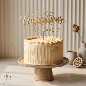 Op de afbeelding: Een goudkleurige taarttopper met het woord Congratulations in een cursief lettertype, geplaatst op een ronde, lichtbeige taart met verticale textuur en gedetailleerde randen, rustend op een bijpassende taartstandaard.