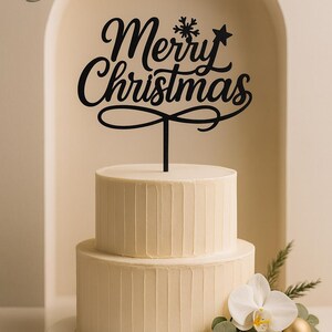 Può includere: Una torta a due piani con glassa bianca e creste verticali. Un cake topper nero recita "Merry Christmas" con un design a fiocco di neve e stella. La torta è decorata con orchidee bianche, verde e un ornamento dorato.