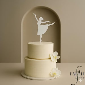 Op de afbeelding: Een crème-kleurige taart met twee lagen, met een gouden band en witte bloemen. Een witte ballerina taarttopper is in het midden geplaatst. De taart staat op een witte taartstandaard. De achtergrond is neutraal.