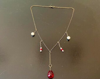 May include: A delicate gold-toned necklace with a teardrop-shaped, red pendant. The necklace features small white and red beads and a simple chain design. A stylish piece of jewelry.