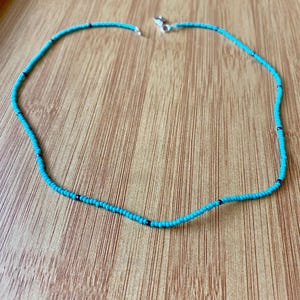 May include: A delicate beaded necklace featuring small turquoise beads with interspersed black beads. The necklace is secured with a silver-colored clasp. The necklace is displayed on a wooden surface.