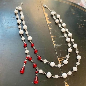 May include: A double-strand necklace featuring white spherical beads and translucent clear beads interspersed with vibrant red spherical beads. The necklace includes two dangling red teardrop-shaped pendants attached to silver-toned metal chains.