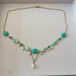 May include: A gold-tone chain necklace featuring turquoise-colored oval beads with a textured crosshatch pattern, small green accent beads, and tiny white pearls. The center of the necklace displays a V-shaped drop with a single white pearl pendant.