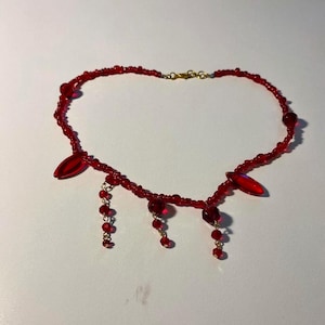 May include: A red beaded necklace featuring a series of small round red beads along the main strand, accented with two larger marquise-shaped red beads and three dangling charms composed of small red beads connected by thin gold-toned metal links.