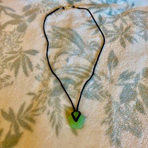 May include: A necklace featuring a black cord and a light green, irregularly shaped pendant. The pendant is attached to the cord with a black V-shaped connector. The necklace has a gold-colored clasp.