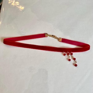 May include: A red velvet choker necklace with gold-colored chain and clasp. The necklace features three dangling strands of small, red beads. The choker is displayed on a white surface.