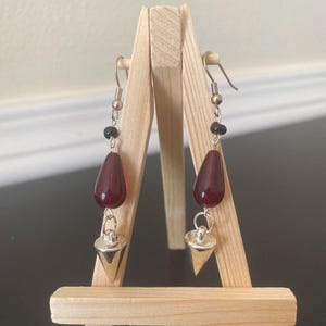 May include: A pair of dangle earrings with a dark red teardrop-shaped bead, a small black bead, and a silver-colored, triangular charm. The earrings are displayed on a wooden easel.
