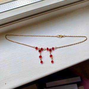 May include: A delicate gold-colored necklace featuring a series of small, vibrant red beads. The necklace has a simple chain design with a clasp closure. Two strands of red beads hang from the center, adding a unique touch to the jewelry.