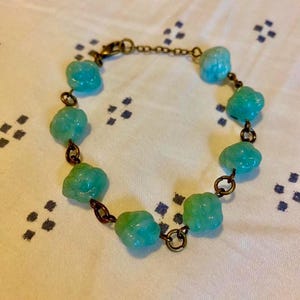 Puede incluir: Una pulsera con cuentas en forma de flor de color azul claro y verde. Las cuentas están conectadas por pequeños eslabones y una cadena de color bronce oscuro. La pulsera tiene un cierre de broche. La joya se exhibe sobre una superficie blanca con un patrón azul.