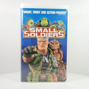May include: A VHS tape case for the movie Small Soldiers with a blue background. The cover features a central human-like soldier figure in camouflage, a creature with a large eye, a green monster, and a brown ape-like character with text.