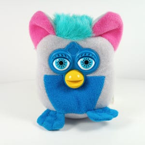 May include: A vintage Furby plush toy with a gray body, blue accents, and pink ears. The toy has large blue eyes, a yellow beak, and a tuft of turquoise fur on its head. The Furby is a collectible electronic toy from the 1990s.
