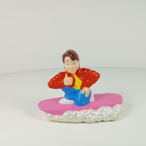 May include: A plastic figurine of a character with brown hair, wearing a red jacket, yellow shirt, and blue pants, kneeling on a pink surfboard. The surfboard features a white, textured wave design at the front, giving the appearance of motion.