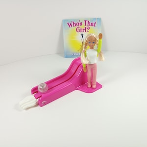 May include: A vintage doll with blonde hair, wearing a white shirt, pink striped pants, and a green sleeve, stands next to a pink plastic slide. A small book titled "Who's That Girl?" is in the background. A clear ball sits on the slide.
