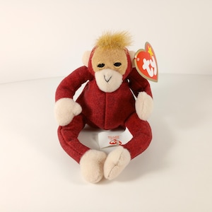 May include: A plush monkey toy in a seated position. The monkey has a red body, tan face, and cream-colored hands and feet. It has a small tuft of tan hair on its head and black eyes. A red heart-shaped tag is attached to the monkey's ear.