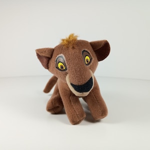 May include: A plush lion cub toy in shades of brown, with a tuft of light brown fur on its head. The toy has large, expressive eyes with yellow irises and black pupils, and a black nose. The cub is in a seated position.