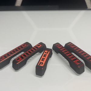 May include: Six black and red Jeep grab handles. The handles have a rectangular shape with a series of parallel slots and the word "JEEP" in orange on one handle. The handles are designed for a vehicle.