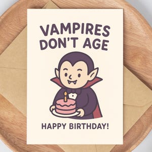 May include: A greeting card with a cartoon vampire holding a pink birthday cake with a lit candle. The card reads "VAMPIRES DON'T AGE HAPPY BIRTHDAY!" The card is on a wooden tray with a brown envelope.