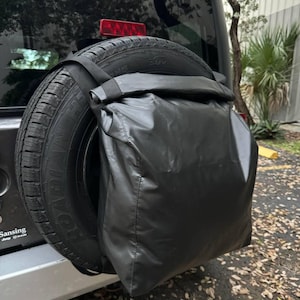 以下が含まれることがあります： Jeepの後部に取り付けられた黒いスペアタイヤバッグ。バッグは耐久性のある防水素材で作られており、黒いストラップで固定されています。タイヤは黒いSUVタイヤです。背景には窓といくつかの葉が含まれています。
