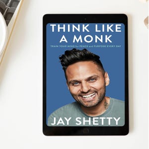 Könnte beinhalten: Ein schwarzes Tablet zeigt das Buchcover von "Think Like a Monk" von Jay Shetty. Das Cover zeigt ein Foto des Autors, der vor blauem Hintergrund lächelt. Titel und Untertitel des Buches sind in weißem Text.