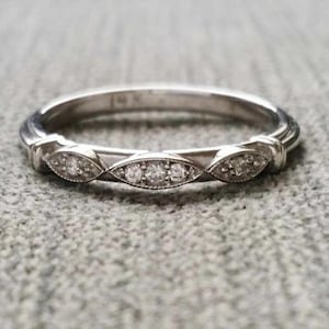 Peut inclure: Une bague en argent au design délicat. La bague présente une série de sertissages en forme de marquise, chacun orné de petits pierres précieuses scintillantes. L'anneau est fin et poli, avec une texture subtile. La bague est posée sur un fond gris.