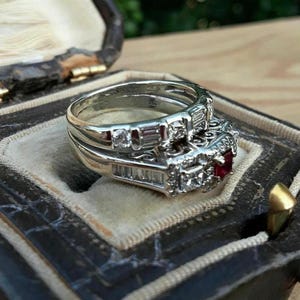 May include: A silver engagement ring set with a ruby and multiple diamonds, resting in a vintage jewelry box. The ring features a central ruby stone and is surrounded by smaller diamonds. The box is dark with a velvet interior.