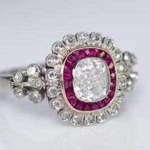 May include: An antique ring featuring a large, square-cut, clear gemstone at the center, surrounded by a halo of smaller, clear stones and a border of red gemstones. The ring has a silver band and intricate detailing.