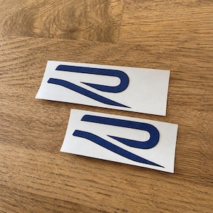 Könnte beinhalten: Zwei identische dunkelblaue Aufkleber mit einem stilisierten Buchstaben R. Jeder Aufkleber ist auf einem rechteckigen weißen Trägerpapier gedruckt und zeigt eine schlanke, längliche und geschwungene Schriftart mit matter Oberfläche.
