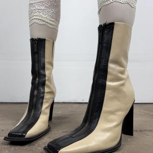 Puede incluir: Un par de botines con diseño bicolor en paneles de cuero color crema y negro. Los botines cuentan con punta cuadrada, tacón de bloque alto y una cremallera negra prominente que recorre verticalmente el centro frontal de cada caña.