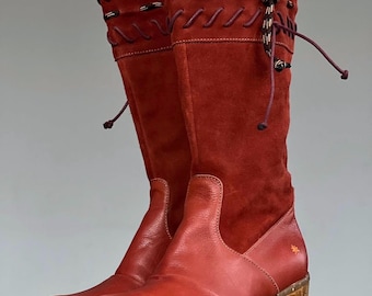 May include: A pair of tall, reddish-brown boots with a suede upper and leather lower. The boots have a wooden heel and decorative stitching and lacing. The boots are held up by a hand.
