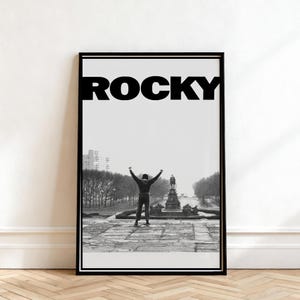 May include: Black and white framed poster featuring the word "ROCKY" in bold letters above an image of a person with arms raised in triumph. The image is a scene from the movie, with a statue and fountain in the background.