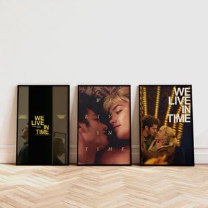 以下が含まれることがあります： 3つの額入り映画ポスター。ポスターには、白い文字で「WE LIVE IN TIME」というタイトルが書かれています。最初のポスターは黒と金のデザインです。2番目のポスターはキスをしている2人を示しています。3番目のポスターは抱き合っている2人を示しています。