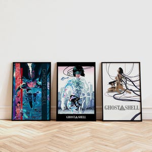 Puede incluir: Tres láminas enmarcadas de arte de Ghost in the Shell. Las láminas presentan ilustraciones de estilo anime de personajes y escenas de la serie. La paleta de colores incluye azules, rojos y blancos. El texto "Ghost in the Shell" es visible.