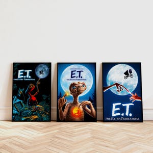 May include: Three framed movie posters featuring the film "E.T. the Extra-Terrestrial." Each poster has a black frame and showcases iconic scenes with the E.T. character against a moonlit backdrop. The posters are displayed on a wooden floor.