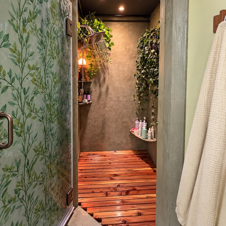 May include: A shower stall featuring a slatted cedar wood floor, gray concrete walls, and a glass door with a green leaf pattern. A square shower head sprays water, surrounded by hanging green plants and shelves holding various bottles.