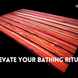 May include: A rectangular bath mat constructed from parallel slats of reddish-brown wood with visible grain patterns. The text VIDA appears in the top right corner, and the text ELEVATE YOUR BATHING RITUAL is printed in white at the bottom.