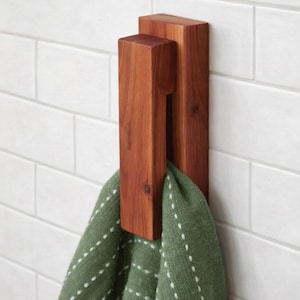 May include: A wooden towel hook in a rectangular design. The hook is made of reddish-brown wood and is mounted on a white tiled wall. A green towel with white dotted lines is hanging from the hook.